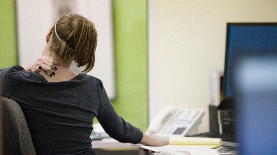 A seated person touching the back of the neck while facing a desktop computer.